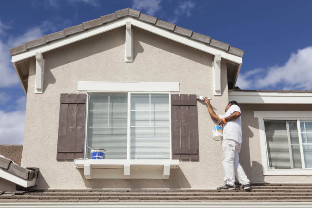 Bright house exterior being painted with brown shutters, professional painter applying fresh paint on the wall, blue sky with few clouds, showcasing high-quality exterior painting services.