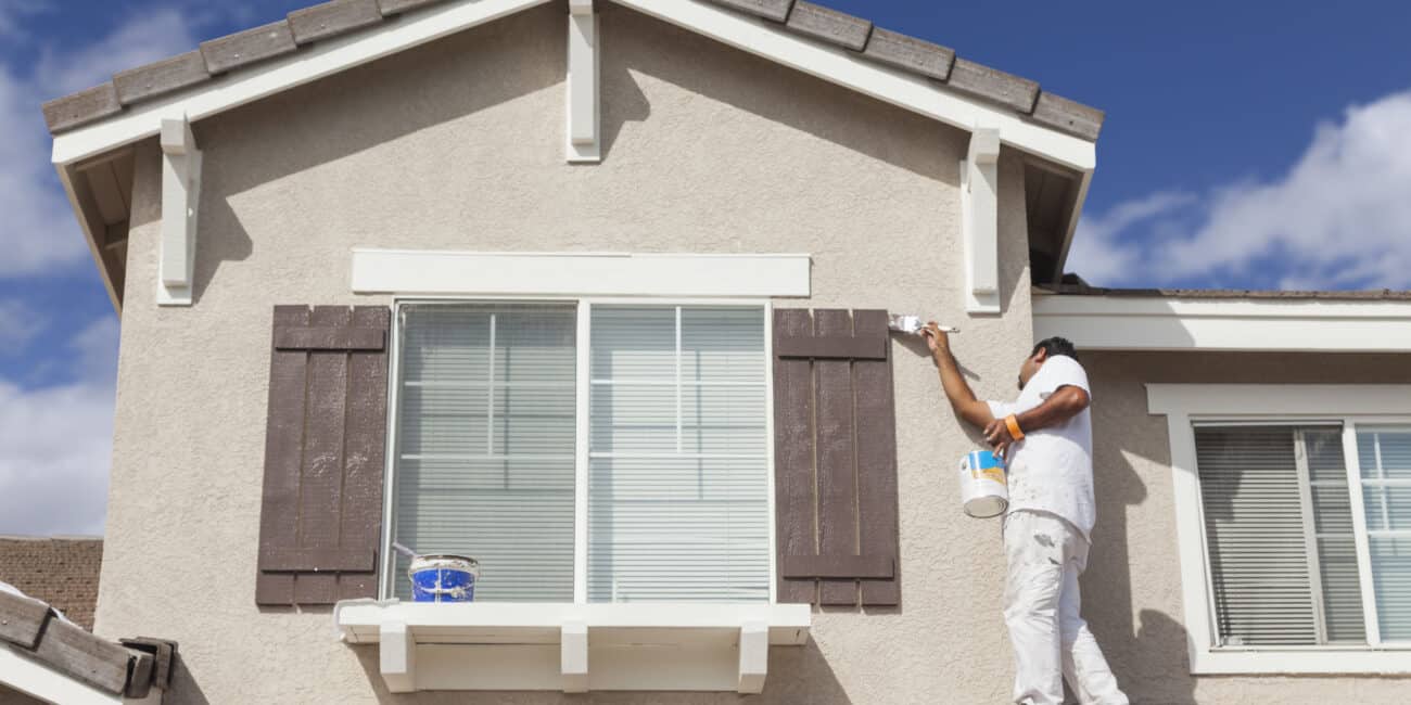 Bright house exterior being painted with brown shutters, professional painter applying fresh paint on the wall, blue sky with few clouds, showcasing high-quality exterior painting services.