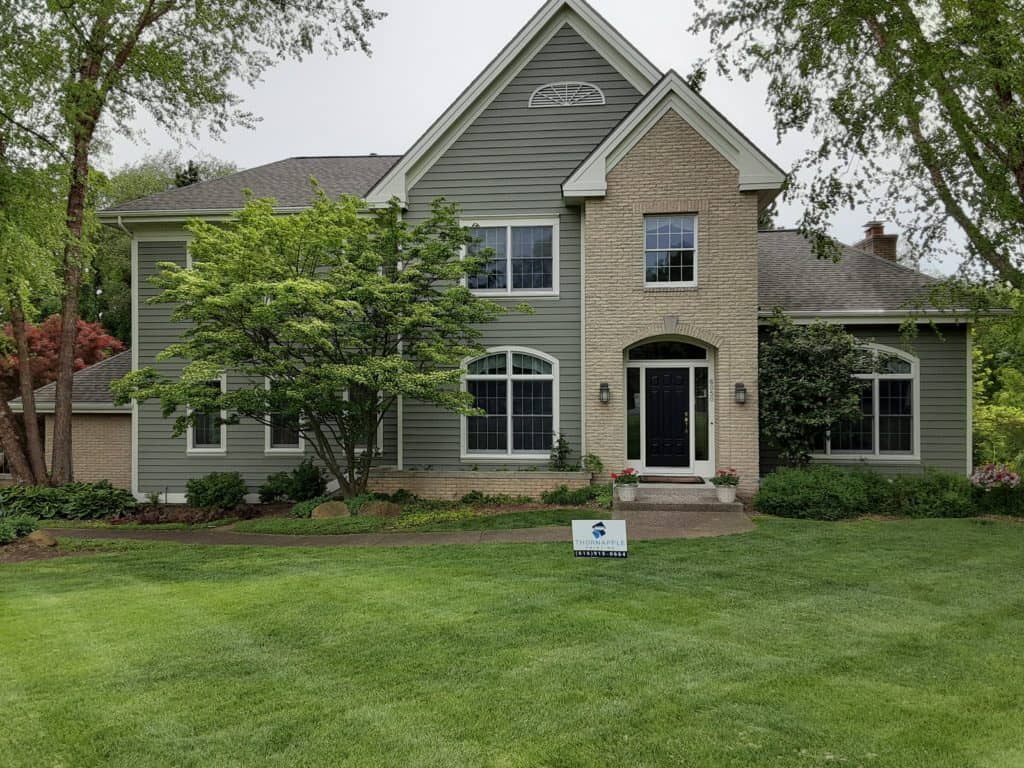 Spacious residential house with modern green siding and brick accents, surrounded by lush landscaping and mature trees, showcasing professional exterior painting services.