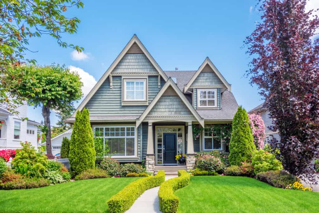 Elegant Craftsman-style house with vibrant landscaping, lush green lawn, colorful flowers, and mature trees, showcasing expert exterior painting and home improvement services by Thornapple Painting.